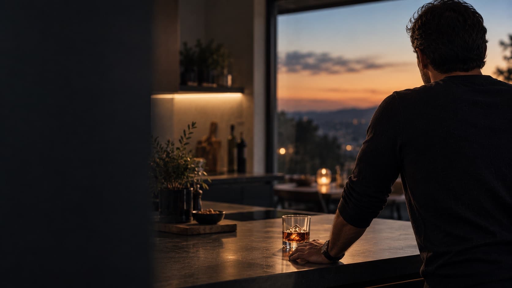 A man standing at a kitchen counter at dusk, looking out over the city, an untouched glass of whiskey beside his hand.